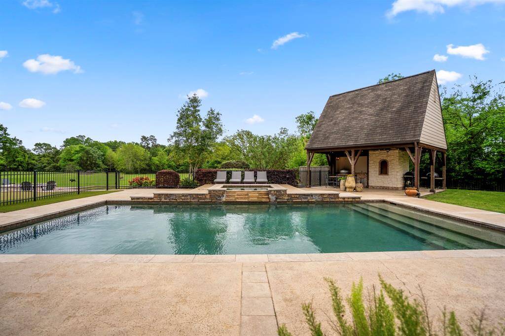 Large swimming pool with a stone deck, surrounded by lush greenery. A charming poolside pavilion offers a shaded area, ideal for relaxing or entertaining.