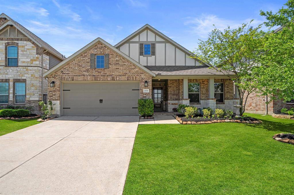 The exterior features a majestic brick and wood ranch-style facade with a well-manicured lawn and a covered patio. The property includes an attached garage with a charming suburban garden with grass and decorative plants and trees.