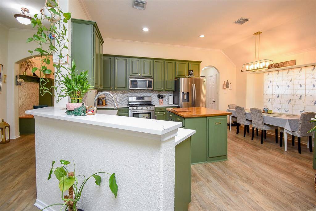 This photo showcases a bright kitchen with green cabinetry, stainless steel appliances, and a wood-topped island. The adjacent dining area features a large table under a modern light fixture. The space has warm wood floors creating a welcoming atmosphere.