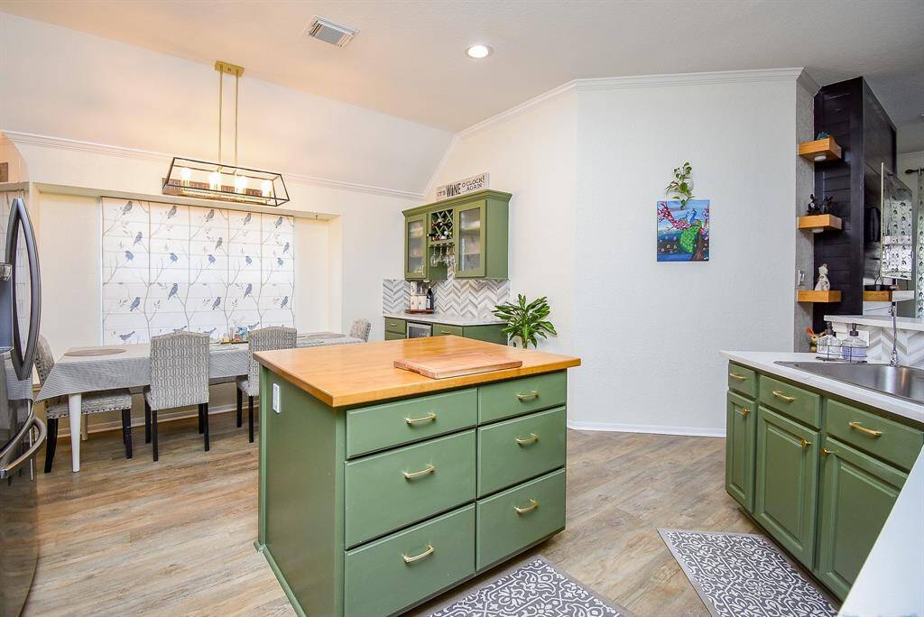 This kitchen features a charming blend of green cabinetry with brass handles and a central island with a wooden countertop. The breakfast area is well-lit. The space is open and bright with modern fixtures and light wood flooring, creating a cozy and inviting atmosphere.