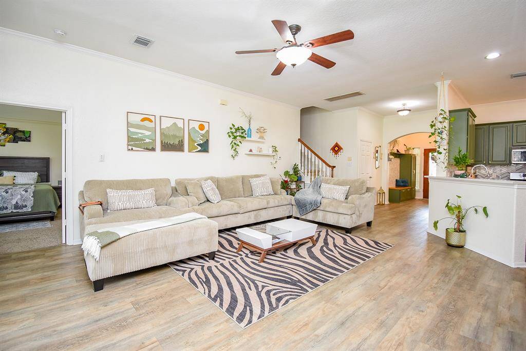 Spacious and bright living area. The room connects to an open kitchen and there's a visible stairway leading upstairs. A ceiling fan and natural light enhance the welcoming atmosphere.