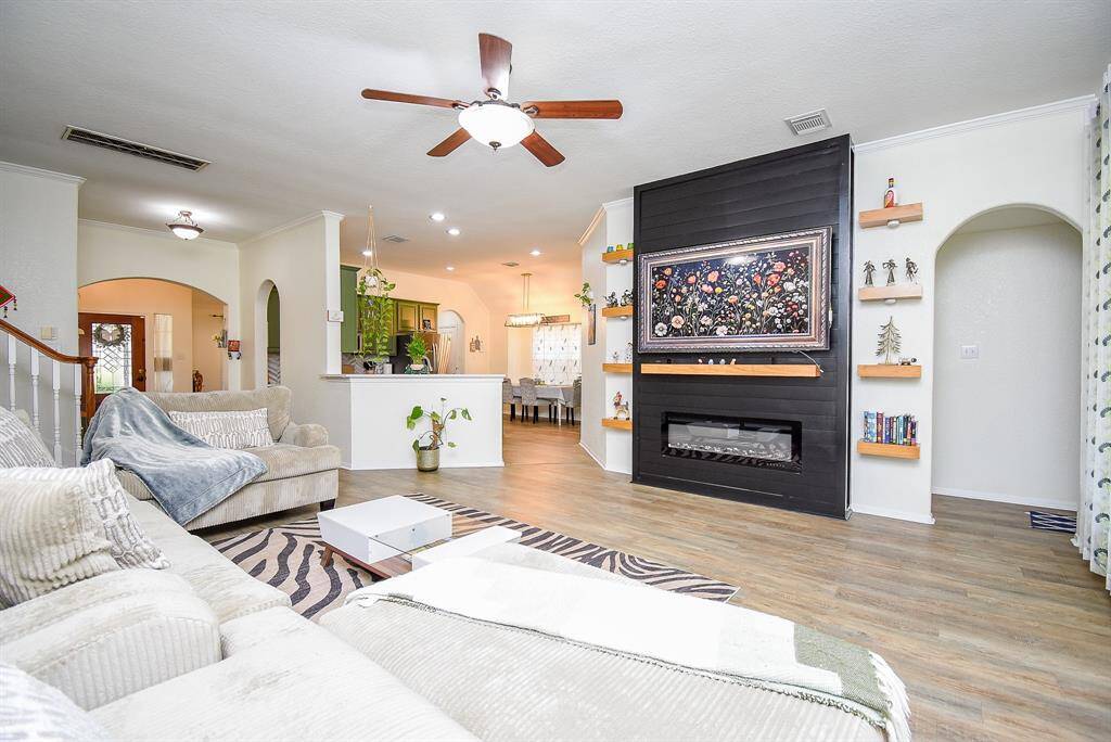 Bright and spacious living area featuring modern wood flooring, an elegant fireplace, and a ceiling fan. The open layout flows into a dining space and kitchen, creating a warm and inviting atmosphere. Perfect for comfort and entertaining.