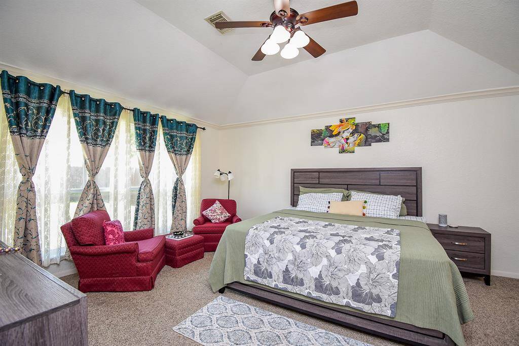 The primary bedroom features a vaulted ceiling and a ceiling fan, offering a spacious feel. It includes a large window with elegant drapes, allowing natural light to fill the room. The space has a cozy sitting area.