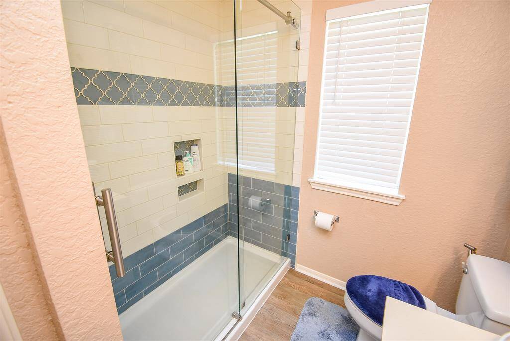This bathroom features a modern glass-enclosed shower with stylish blue and white tile accents, a convenient built-in shelf, and a nearby window providing natural light.