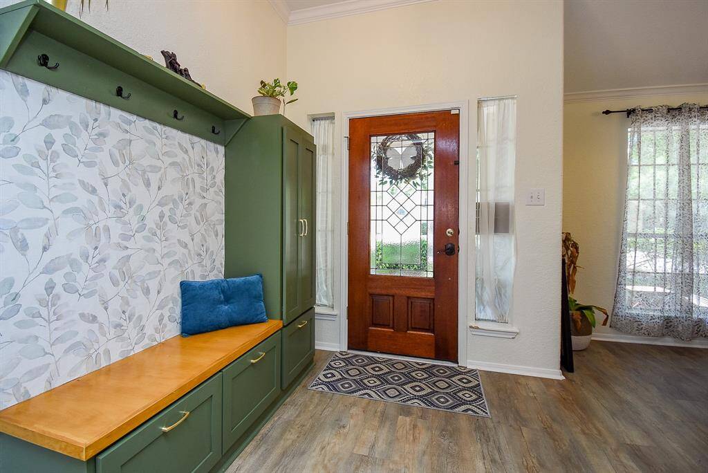 This welcoming entryway features a wooden front door with decorative glass panels, flanked by tall side windows. A built-in green storage bench with a patterned backsplash and hooks offers functionality and style. The space is adorned with light-colored walls and wood-style flooring, creating a cozy yet elegant atmosphere.