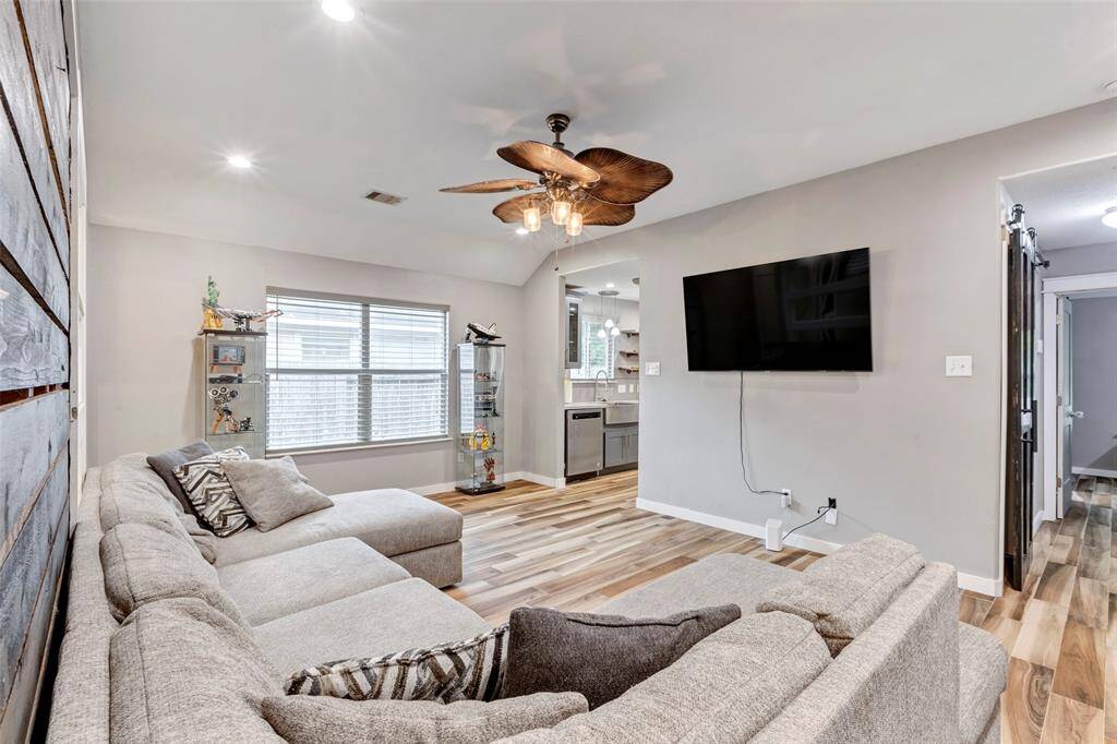 A wide view from the corner of the living room captures the full space, with a peek into the kitchen through a doorway and a hallway leading to additional bedrooms and versatile living areas.