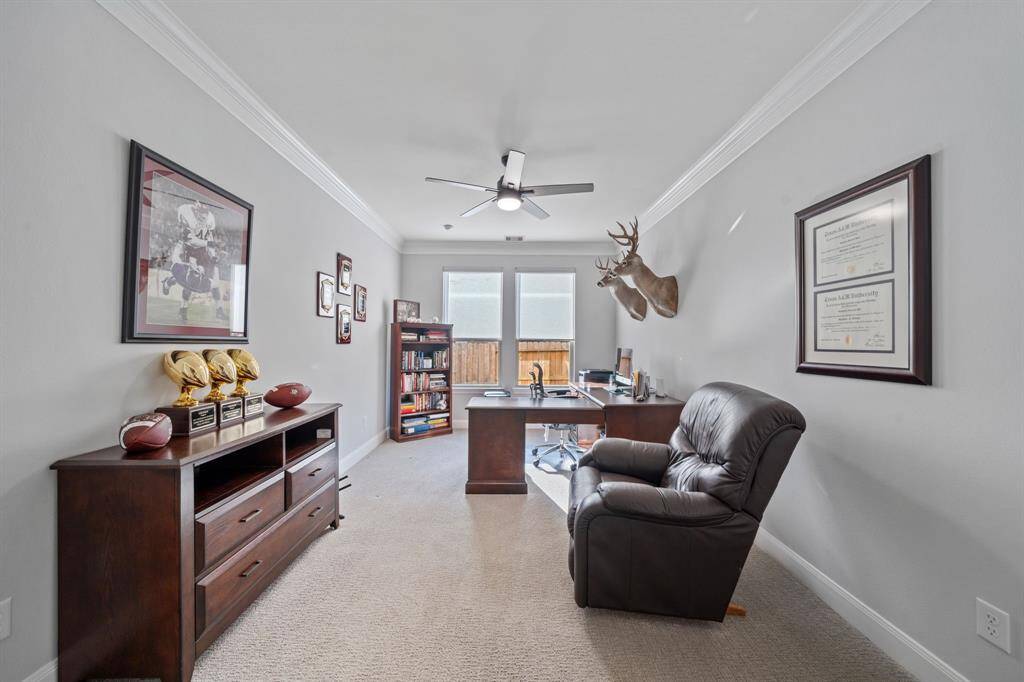 The office features elegant glass doors and plush carpeting, creating a comfortable and stylish workspace. Crown molding, a ceiling fan, and roller shades add functional comfort.
