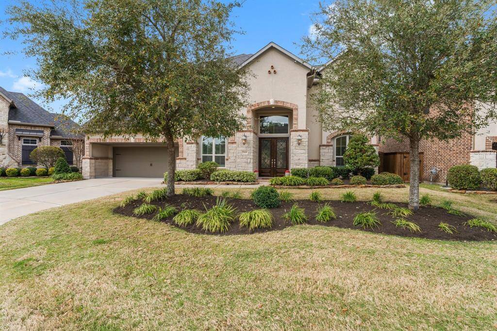 This photo showcases a charming, well-maintained home with a stone and brick exterior. The front yard is landscaped with lush greenery and trees. The property features a spacious driveway leading to a 3-car garage.