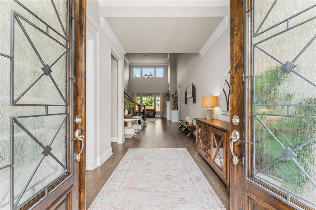 This inviting entryway features elegant glass double doors opening to a bright, spacious hallway with stylish wood flooring. The open layout extends to a cozy living space with large windows, creating a welcoming and airy atmosphere.