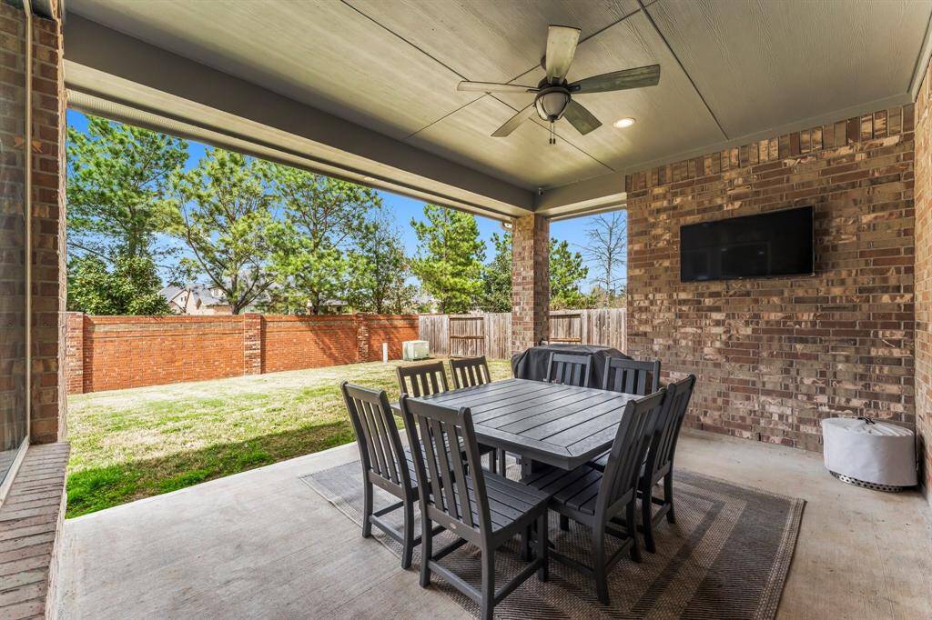 This photo showcases a spacious covered patio with a ceiling fan, outdoor dining set, and wall-mounted TV, overlooking a well-maintained backyard with a brick privacy wall.