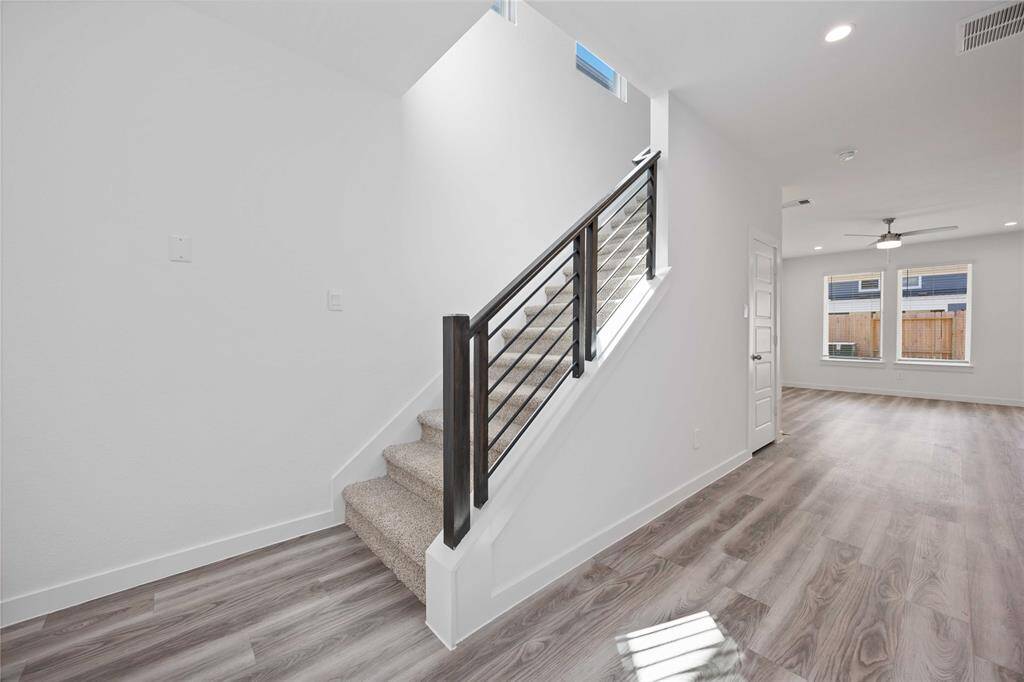 This photo showcases a modern interior with an open layout, featuring light-colored wood flooring and a staircase with sleek black railings. The space is bright with natural light from large windows, and there's a ceiling fan in the living area.
