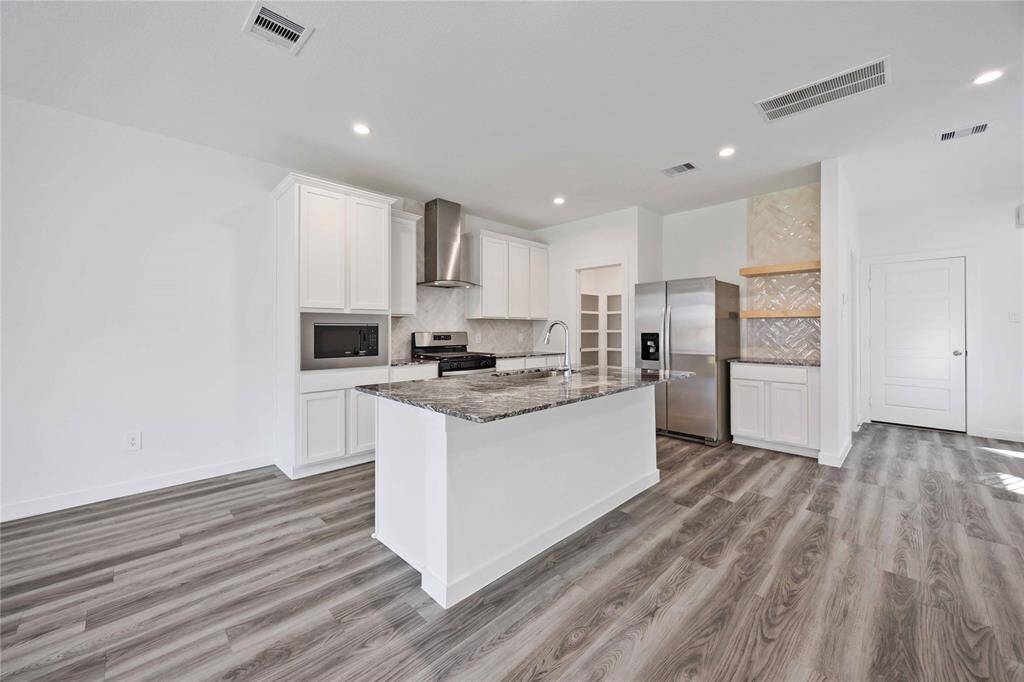 This modern kitchen features sleek white cabinetry, granite countertops, and stainless steel appliances. It includes a spacious island with a sink, stylish backsplash, and ample storage with a pantry and built-in microwave. The open layout and wood-look flooring enhance its contemporary appeal.