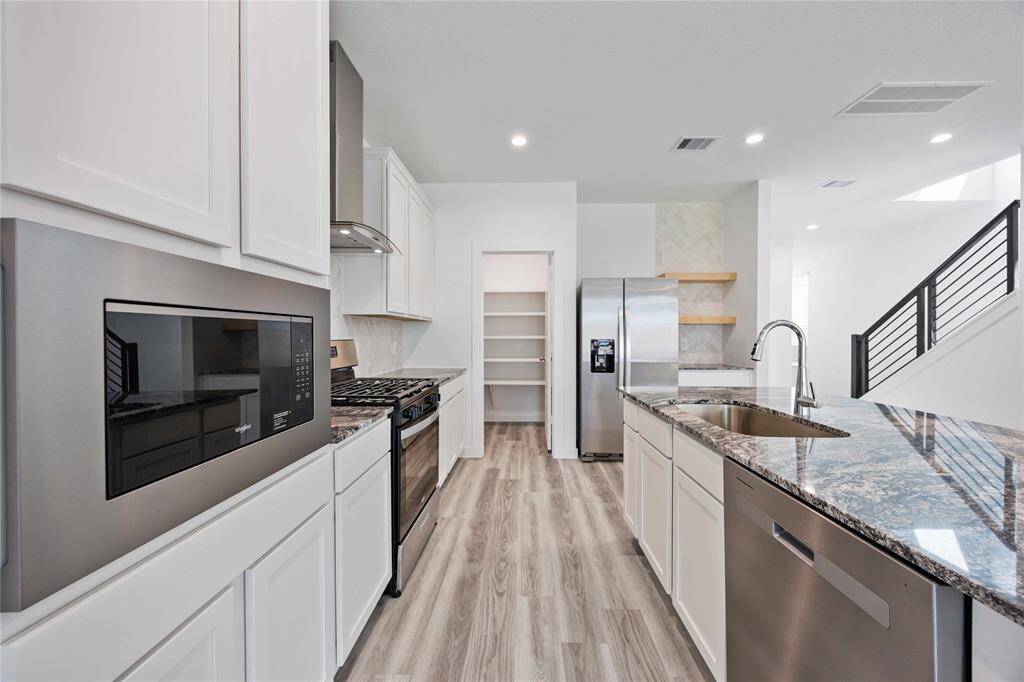This modern kitchen features sleek stainless steel appliances, white cabinetry, and a stylish granite countertop. The open layout includes a spacious pantry, a center island with a sink, and wood-look flooring, creating a bright and functional space perfect for cooking and entertaining.