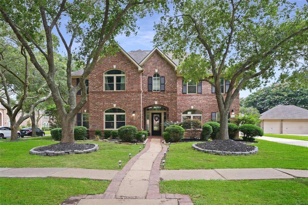 Charming two-story brick home with large windows, elegant landscaping, and mature trees. A welcoming pathway leads to the front door, set in a peaceful suburban neighborhood.
