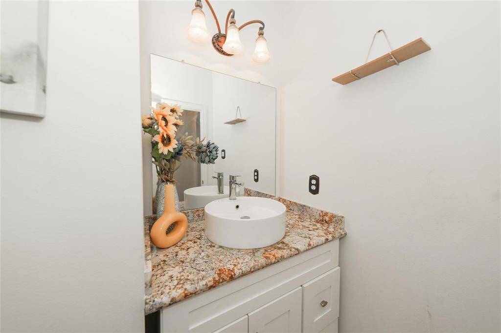 This is a modern bathroom with a granite countertop, a round vessel sink, and a decorative light fixture above. The space has a clean, minimalist look with a vase of flowers adding a touch of color.