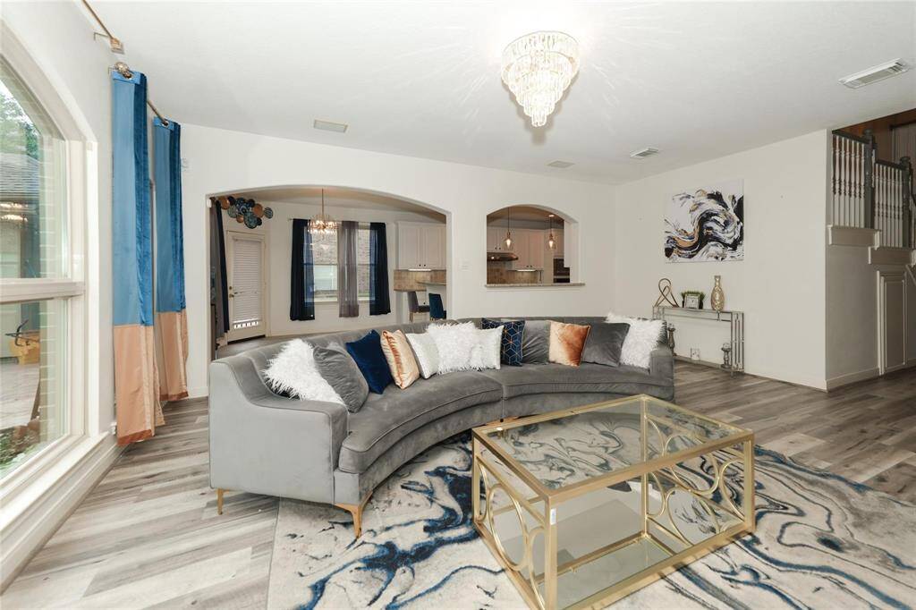 This elegant living room features a large, curved sofa with plush pillows, a stylish glass coffee table, and modern decor. The space is brightened by large windows and a striking chandelier, with an open view to the kitchen and dining area. Perfect for entertaining.