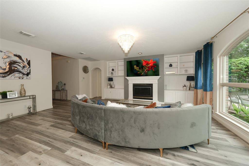 This spacious living room features elegant wood flooring, a cozy fireplace with built-in shelving, and a large, curved sectional. Natural light floods through the expansive window, highlighting the modern decor and open design.