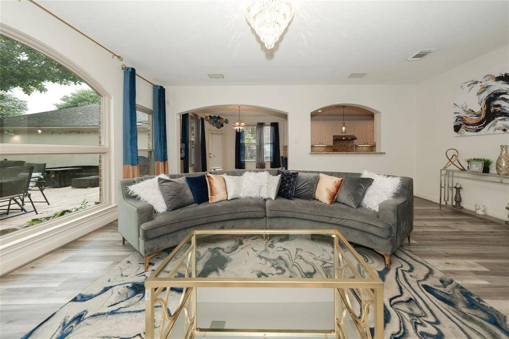 Bright, open living room with a large window overlooking the patio. Features a stylish curved sofa, modern decor, and a gold-accented coffee table on a marble-patterned rug. Elegant archways lead to the dining area and kitchen.