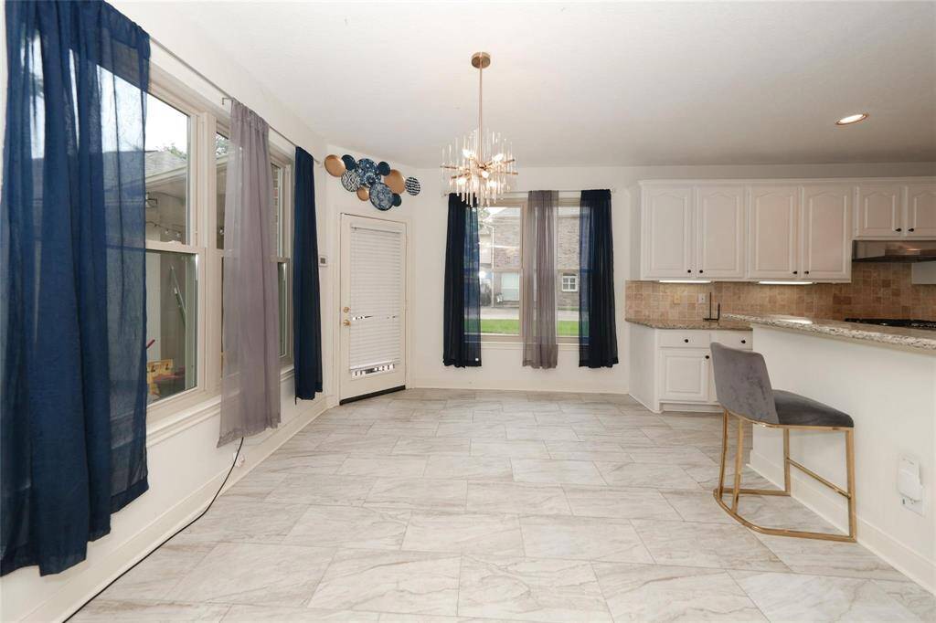 Bright kitchen and dining area with large windows and elegant tile flooring. Features white cabinetry, granite countertops, and a modern chandelier. Cozy breakfast bar with seating.