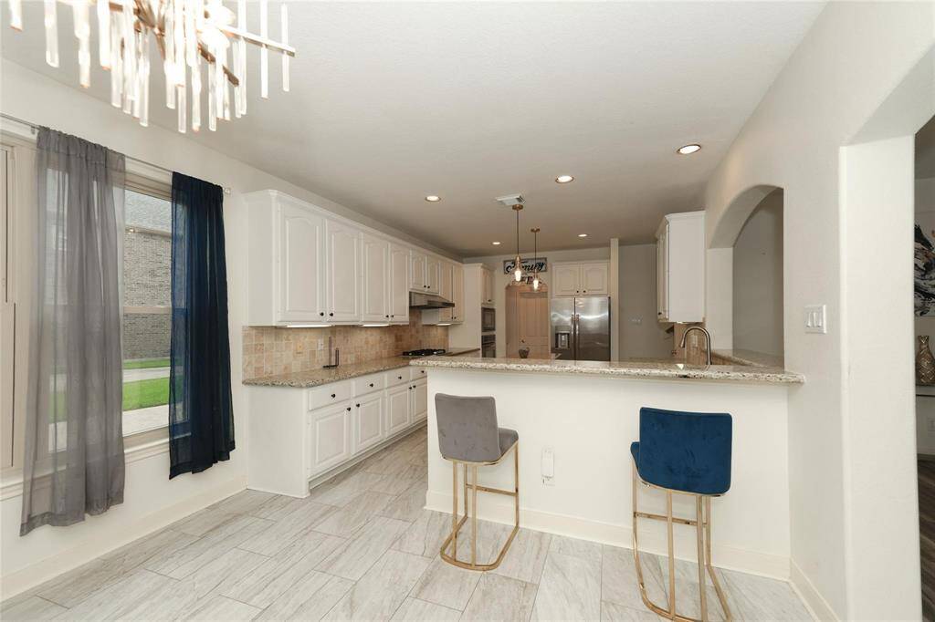 Bright, modern kitchen with white cabinets, granite countertops, and tile flooring. Includes a breakfast bar with stylish seating, stainless steel appliances, and ample natural light from a large window.