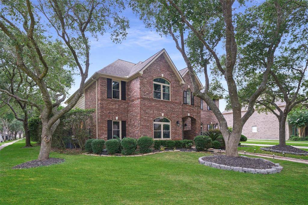 This is a charming two-story brick home with a well-maintained lawn and mature trees. The property features large windows, manicured landscaping, and a corner lot location.