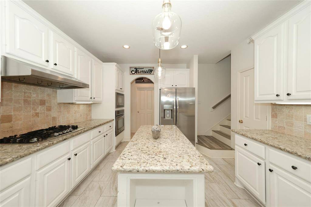 This kitchen features modern white cabinetry, granite countertops, and a central island. It includes stainless steel appliances, a gas cooktop, and a cozy tile backsplash. The space is well-lit with recessed lighting and pendant fixtures, offering a welcoming atmosphere.