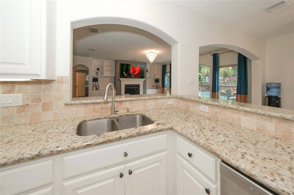 This kitchen features a granite countertop with a dual sink and overlooks an open living area with a fireplace. Large windows offer a view of the backyard and pool, creating a bright and inviting space.