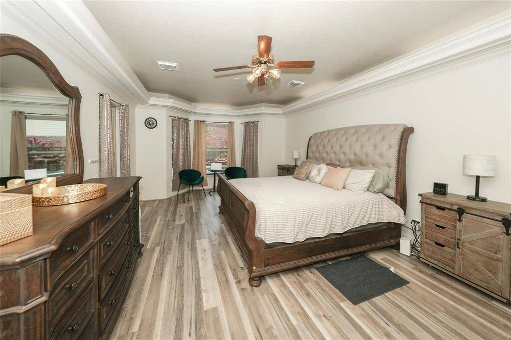 This spacious bedroom features elegant wood flooring and a large, tufted headboard. It includes a ceiling fan, ample natural light from large windows with drapes, and matching wooden furniture, creating a cozy and inviting atmosphere.