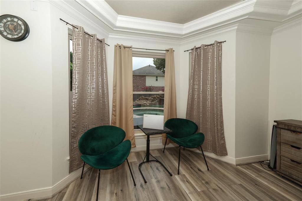A cozy nook with elegant crown molding and large windows overlooking a pool. It features modern flooring, two stylish green chairs, and a small table, perfect for relaxing or reading.