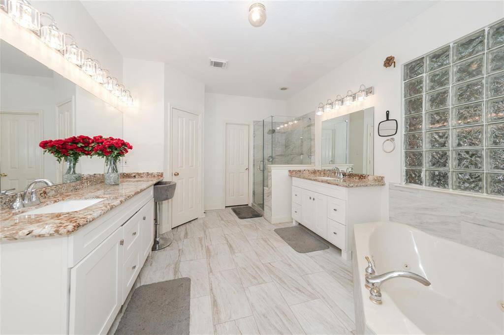 Spacious and elegant bathroom featuring dual sinks with granite countertops, a large soaking tub, a glass-enclosed shower, and bright tile flooring. Natural light streams through a glass block window, enhancing the airy feel.