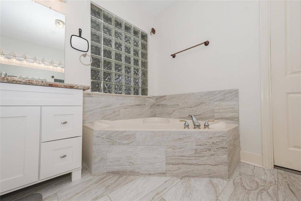 This bathroom features a luxurious corner bathtub with marble-style tiling, a granite countertop with a white vanity, and a glass block wall for added privacy and light.
