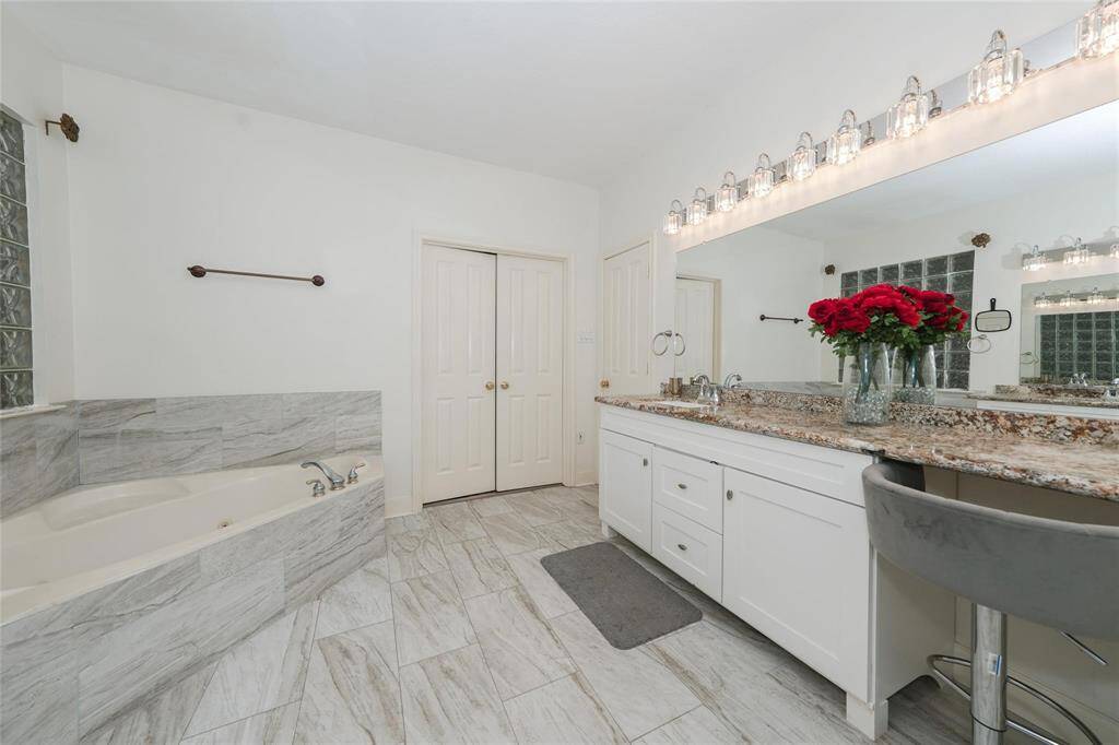 This bathroom features a spacious layout with a large vanity and mirror, granite countertops, and ample lighting. It includes a built-in bathtub with elegant tile and a modern, clean design.