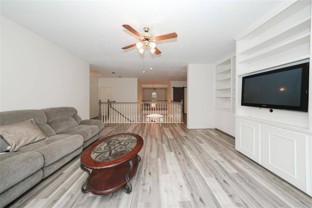 Spacious living area with modern laminate flooring, built-in shelving, and a ceiling fan. Features an entertainment center with a mounted TV and a cozy seating arrangement. Ideal for family gatherings.