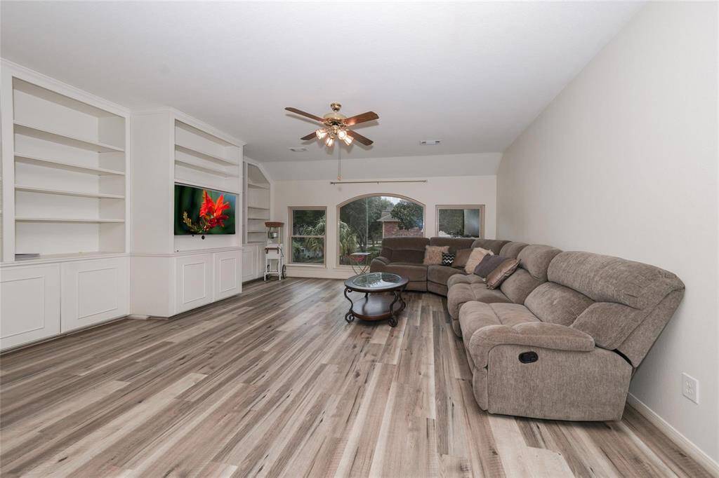 Spacious living room with wood flooring, built-in shelves, a ceiling fan, and a large sectional sofa. Bright with natural light from large windows and features a mounted TV.