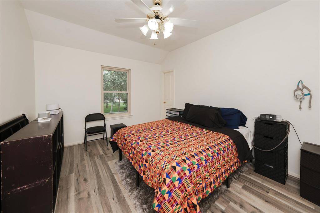 This bedroom features a clean, bright space with white walls, a ceiling fan, and wood-look flooring. It has a window with a view of greenery, a bed with a colorful geometric bedspread, and practical furniture, offering a cozy and functional layout.