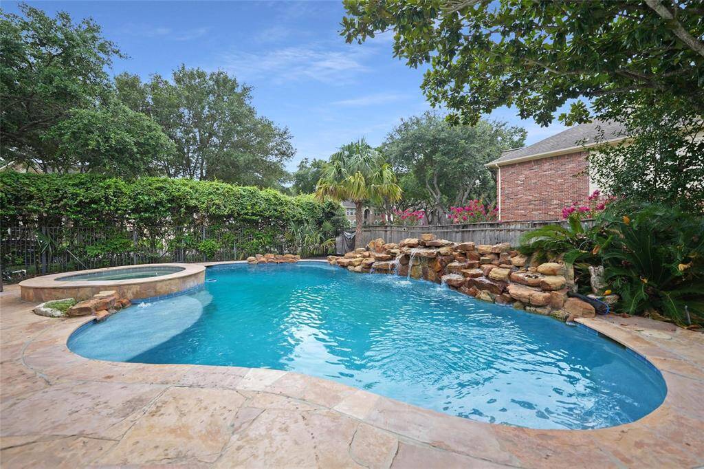 This backyard features a beautiful swimming pool with a stone waterfall and an attached spa, surrounded by lush greenery and a stone patio, perfect for relaxation and entertainment.
