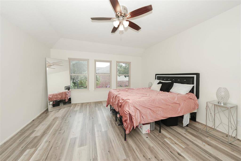 This spacious bedroom features light wood flooring, a ceiling fan, large windows for natural light, and a cozy bed with modern decor.