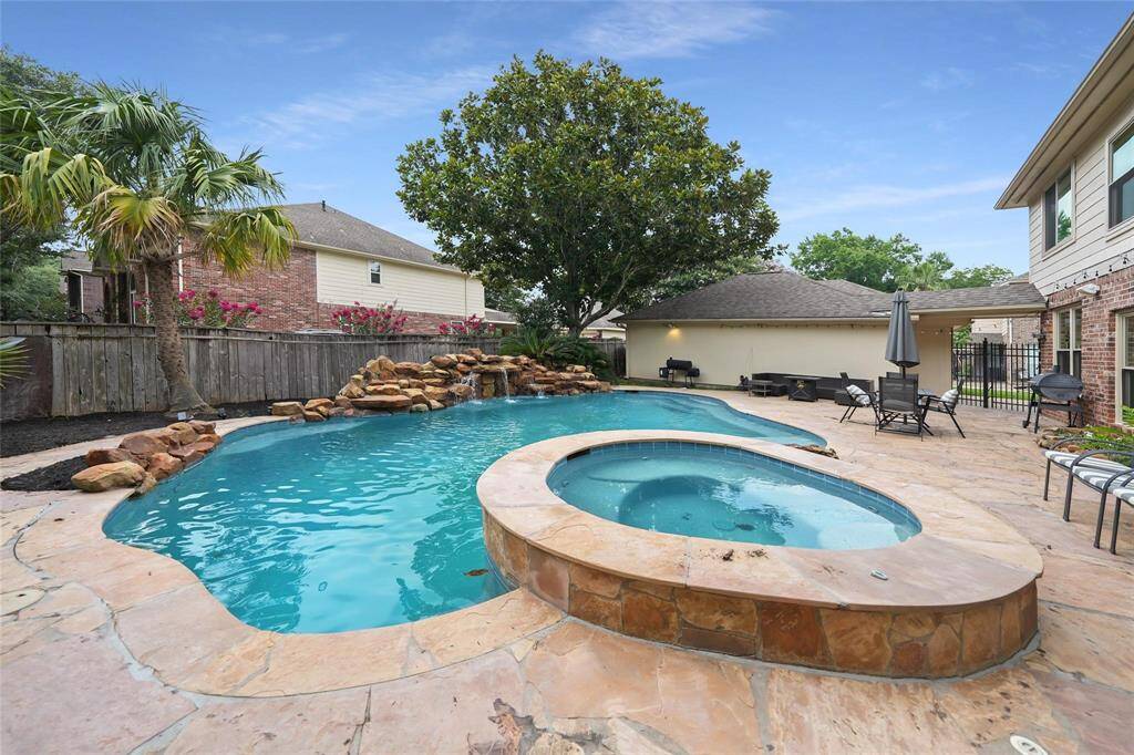 This backyard features a luxurious swimming pool with a rock waterfall and a connected hot tub. Surrounded by a spacious patio with seating areas, it offers a perfect space for relaxation and entertainment. Lush greenery and a privacy fence enhance the serene atmosphere.