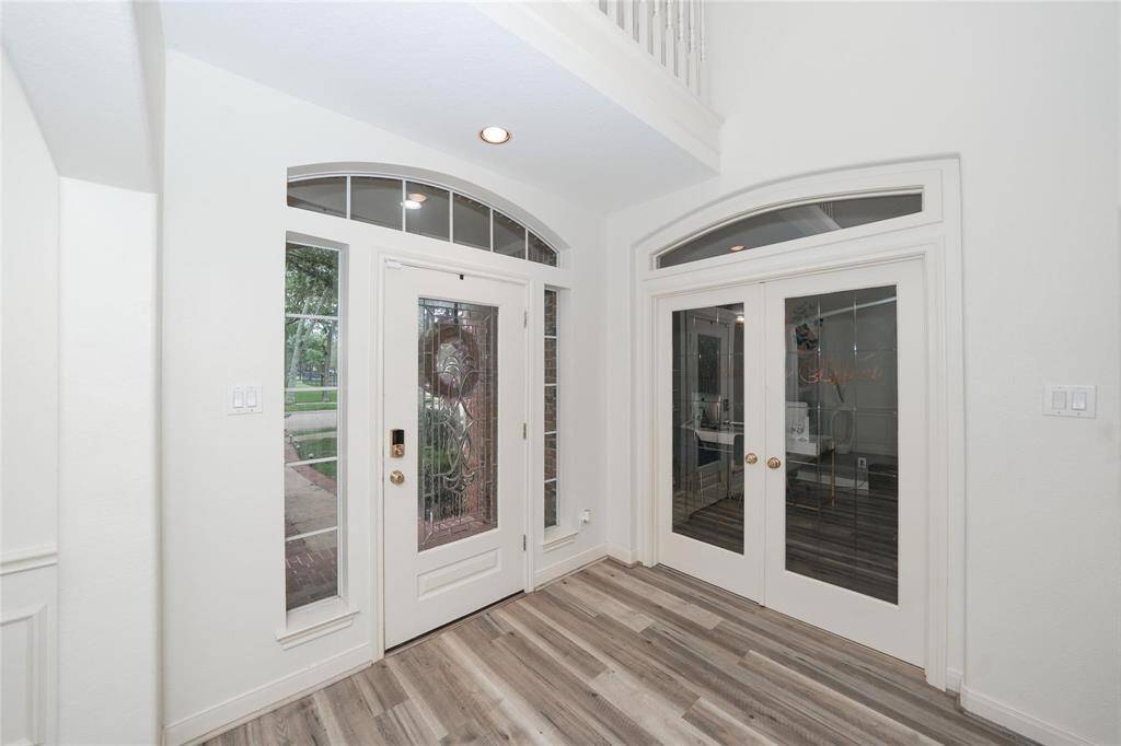 Bright entryway with a high ceiling, featuring a decorative glass front door and side panels. Adjacent double glass doors lead into a room, enhancing the open feel. The space has modern flooring and neutral walls.