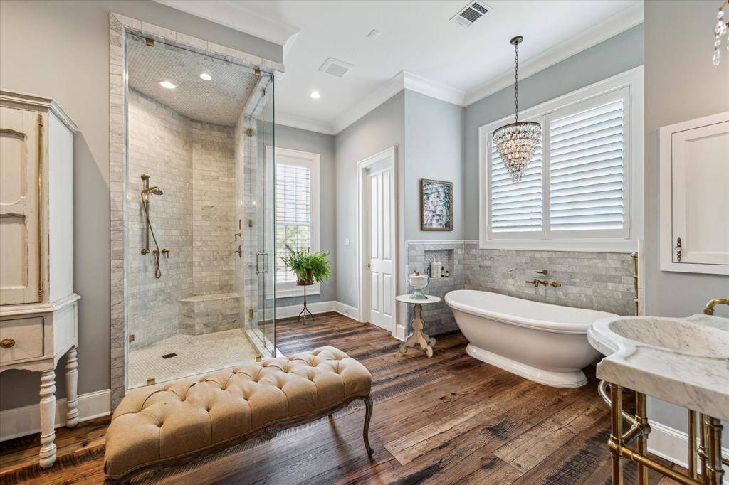 Oversize glass-enclosed steam shower boasts marble tile surround & elegant soaking tub is graced by whimsical chandelier. Windows offer abundant natural light, with plantation shutters added for privacy.