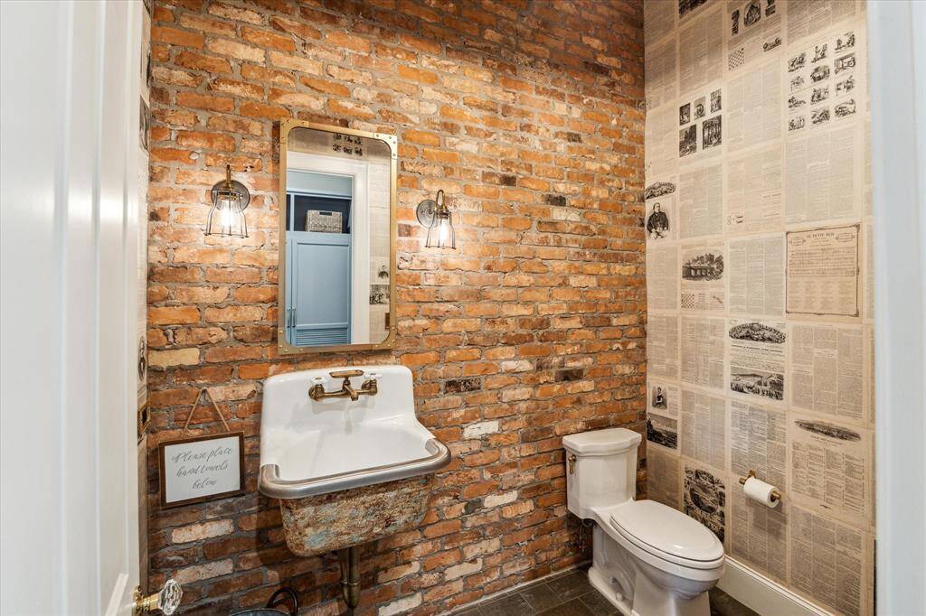 Distinctive utility bath, adjacent to mud room, boasts reclaimed brick accent wall, one-of-a-kind wallpaper fashioned from 1800s vintage French newspapers & custom distressed basin with brass fixtures.