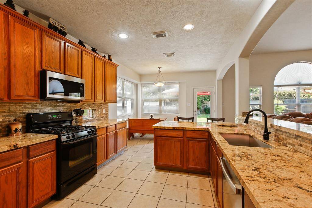 This kitchen features warm wood cabinetry, granite countertops, and a tile backsplash. It includes modern appliances, a gas stove, and a cozy dining nook with natural light from large windows. The space flows into a living area through an arched opening.