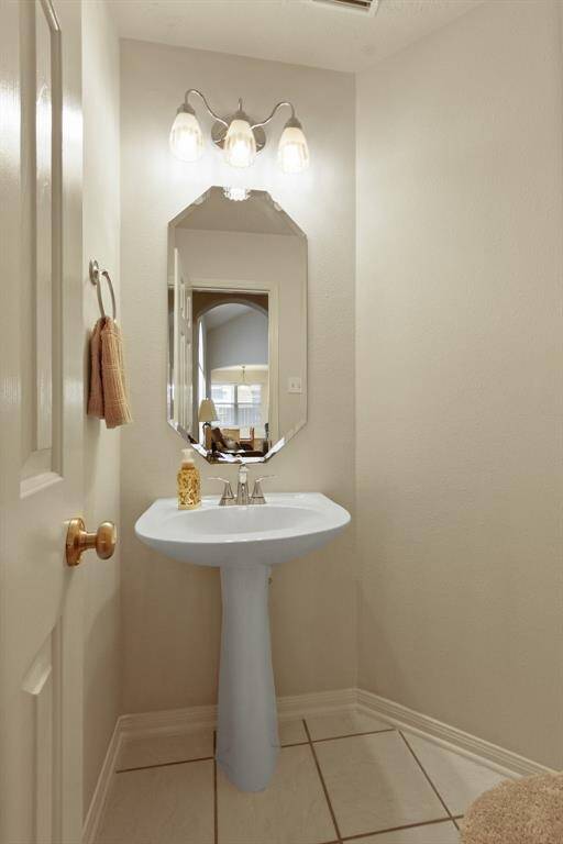 This photo showcases a compact, well-lit bathroom featuring a pedestal sink, a stylish mirror, and a three-light fixture. The neutral tones and tiled floor add to its clean, modern look.