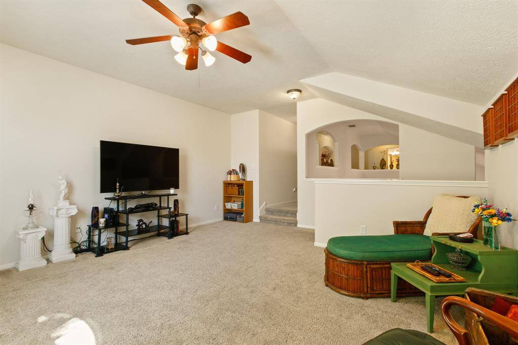 Spacious living / game room with neutral carpet, featuring a ceiling fan. The room has a light, open feel with decorative niches and partial wall divisions, leading into other areas of the home.