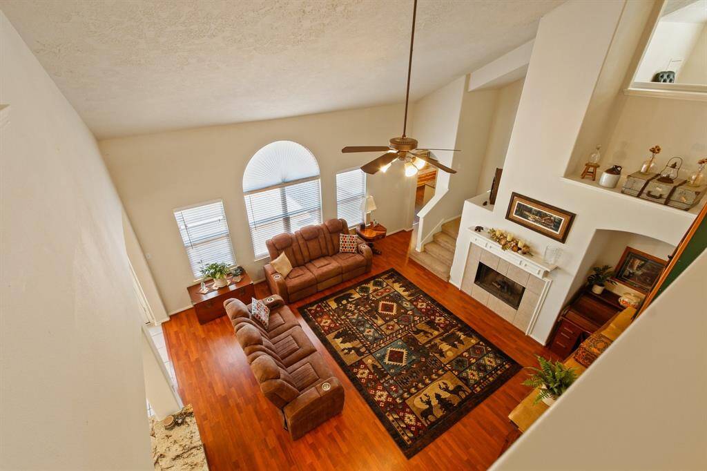 This inviting living room features high ceilings, a ceiling fan, and abundant natural light from large windows. It has hardwood floors, a cozy fireplace, and a comfortable seating area, creating a warm and spacious atmosphere.
