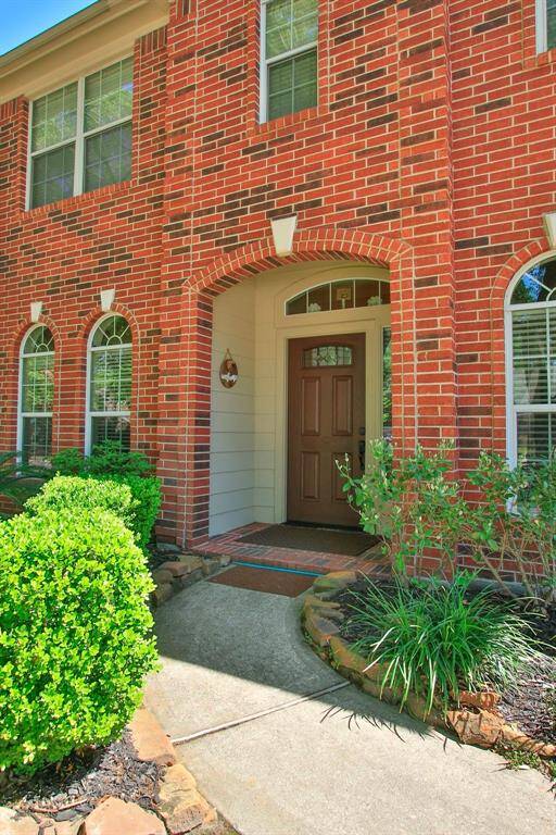 This photo showcases the welcoming entrance of a red-brick home, featuring a covered doorway with a classic wood door. It is flanked by arched windows and surrounded by well-maintained shrubs and a stone-bordered walkway.