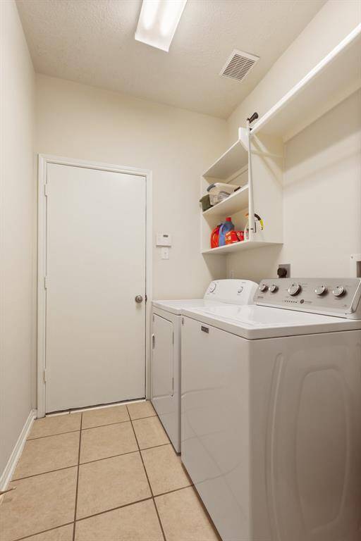 This is a compact laundry room featuring a washer and dryer, with overhead shelving for storage. It has tiled flooring and a door leading to another area of the home.