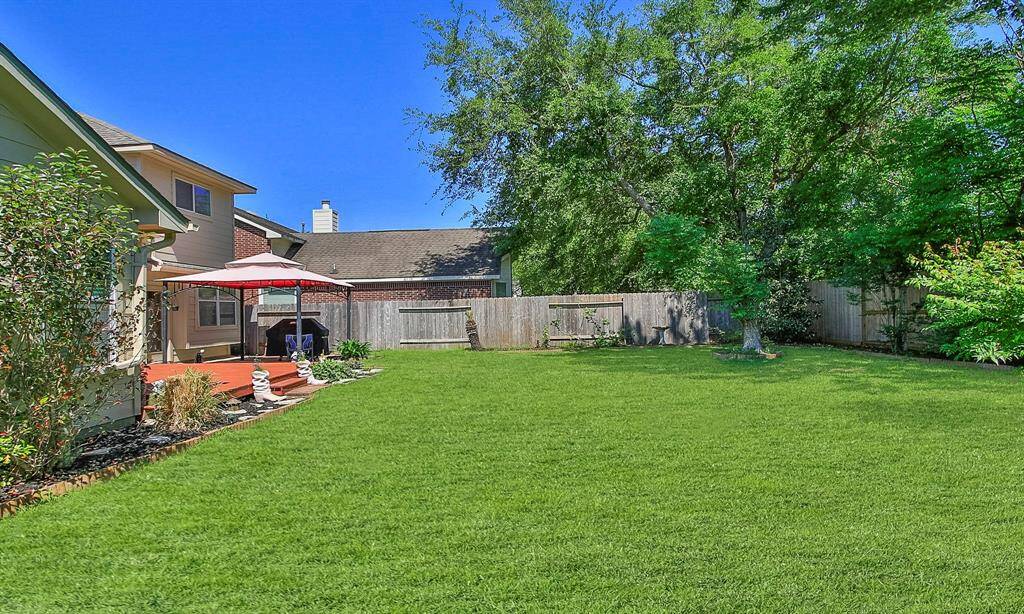 Spacious backyard, bordered by a wooden fence and mature trees. Features a cozy deck with a red umbrella, perfect for outdoor relaxation.