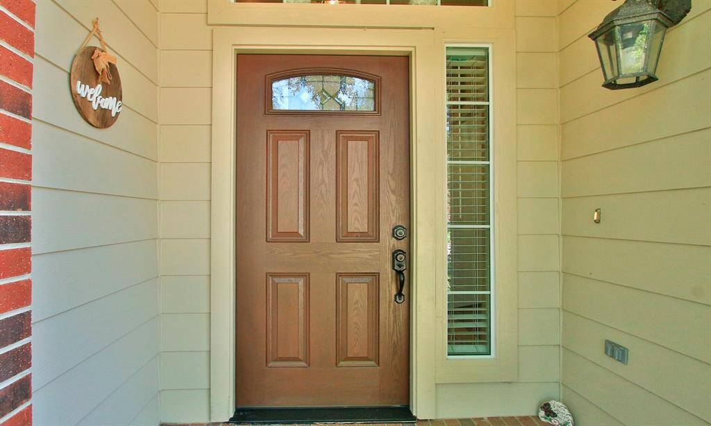 A welcoming front entry with a solid wood door, decorative window accents, and a charming "welcome" sign. The side window with blinds adds natural light, and a classic lantern light fixture enhances the inviting atmosphere.