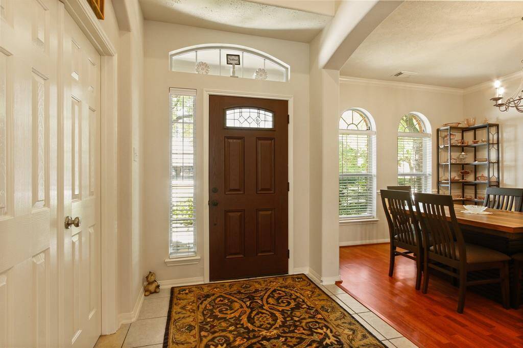 This inviting entryway features a dark wood front door with decorative glass panels, flanked by tall windows for ample natural light. Adjacent is a cozy dining area with warm wood flooring, elegant arched windows, and a stylish chandelier, creating a welcoming atmosphere.